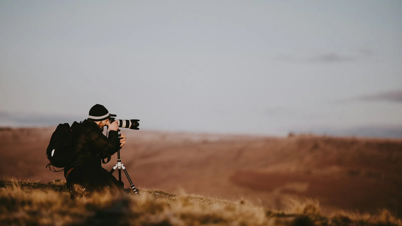 Fotógrafo agachado no campo ao entardecer, mirando a câmera para o horizonte